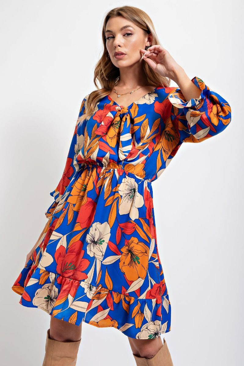 Model wearing a royal blue floral dress with orange, red, and cream flowers, featuring a V-neckline, long sleeves, cinched waist, and tiered skirt.