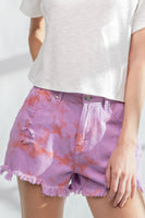 Model wearing lilac and coral tie-dye denim shorts with frayed hem, high-rise waist, and distressed detailing.