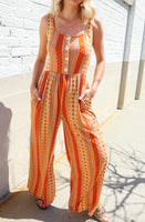 Orange boho wide-leg jumpsuit with button-front bodice, square neckline, and vertical patterned stripes, styled with sandals.